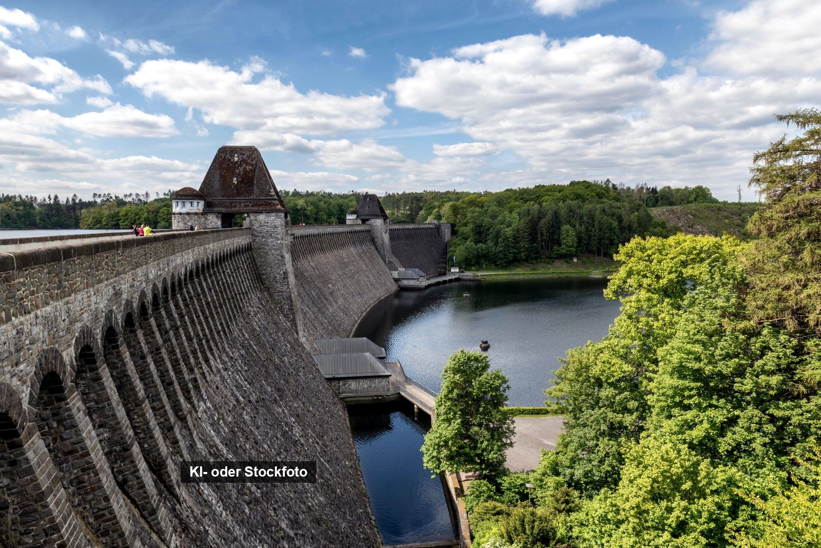 Die Staumauer der Möhnetalsperre mit ihren beiden Türmen, dahinter der Möhnesee und bewaldete Ufer